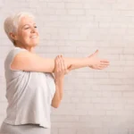 A senior female doing a shoulder stretch indoors.