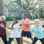 A group of five multi-ethnic seniors taking an exercise class in the park. They are practicing tai chi, standing with their hands raised.