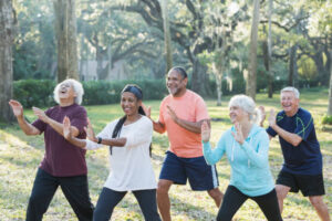 A group of five multi-ethnic seniors taking an exercise class in the park. They are practicing tai chi, standing with their hands raised.
