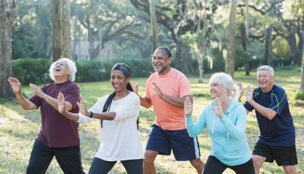 A group of five multi-ethnic seniors taking an exercise class in the park. They are practicing tai chi, standing with their hands raised.