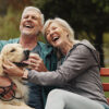 A happy senior couple sitting outside on a bench with their dog.