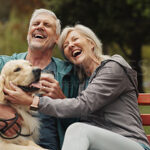 A happy senior couple sitting outside on a bench with their dog.