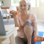 A senior woman is exercising in front of a computer, following a virtual fitness class.