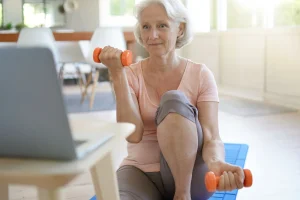 A female senior is doing a virtual fitness session with a computer in front of her.